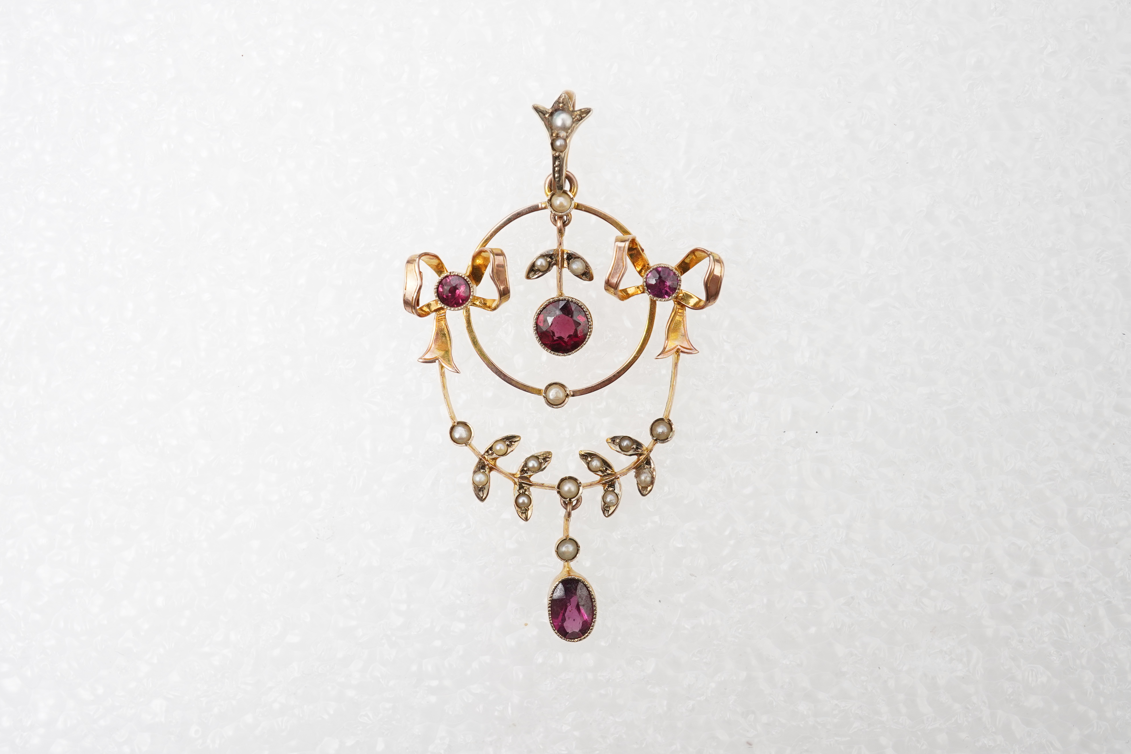 An Edwardian garnet and seed pearl pendant, early 20th century, of foliate design with ribbon bow motifs, set with circular-cut and oval garnets and seed pearls, mounted in gold, length 5.2cm, stamped 9CT, gross weight a
